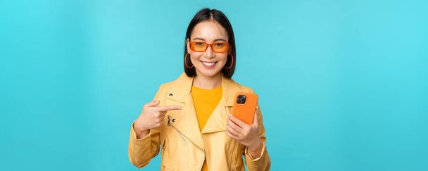 Młoda stylowa azjatycka modelka w modnych okularach przeciwsłonecznych, strój sprężynowy, pokazująca telefon komórkowy, wskazująca na smartfona i uśmiechnięta, stojąca nad niebieskim tłem - Zdjęcie, obraz
