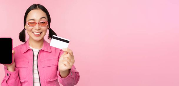 Image of smiling korean woman showing credit card and mobile phone screen, smartphone application interface, paying online, shopping contactless, standing over pink background - Fotoğraf, Görsel