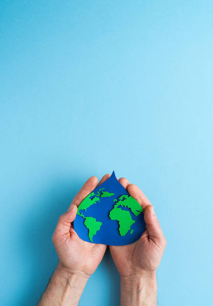 Paper cut out water drop with world map inside on blue background inside palm of hand. Earth day. Vertical photo. - Valokuva, kuva