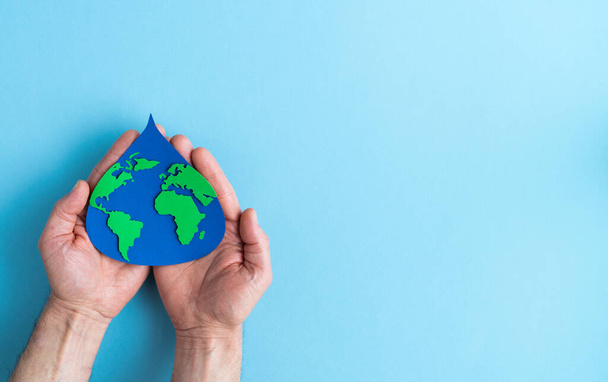 Paper cut out water drop with world map inside on blue background inside palm of hand. Copy space. Earth day. - Photo, image
