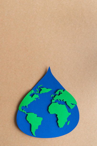 Goccia d'acqua ritagliata su carta blu con mappa del mondo su sfondo cartoncino marrone. Ricevuto. Foto verticale. - Foto, immagini