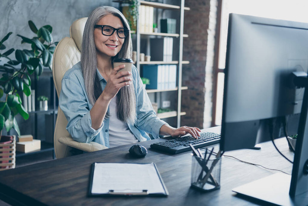 Photo of joyful smile lady ceo pensioner retired sit table drink takeaway mug latte use device monitor prepare project in workspace - Photo, Image