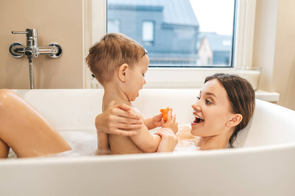 Cheerful mother playing with her child during bathing - Фото, изображение