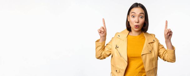 Surprised asian girl pointing fingers up, express interest, showing advertisement ahead, demonstrating promo banner on top, standing against white background - Photo, Image