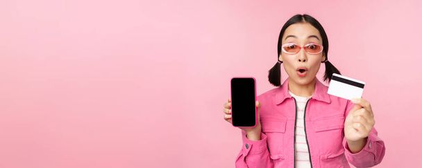 Ázsiai lány mutatja mobiltelefon képernyő és hitelkártya, reagál meglepett kamera, zihálva lenyűgözött, álló felett rózsaszín háttér, online vásárlás koncepció - Fotó, kép
