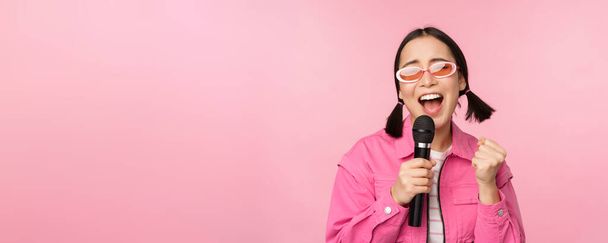 Boldog gyönyörű ázsiai lány énekel mikrofonnal, használata mikrofon, élvezi karaoke, pózol szemben rózsaszín stúdió háttér - Fotó, kép