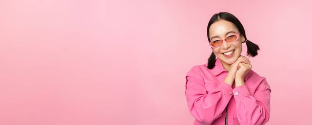Крупный план портрета стильной азиатской женщины в солнечных очках, улыбающейся, выглядящей польщенной, смеющейся кокетливой, стоящей на розовом фоне - Фото, изображение
