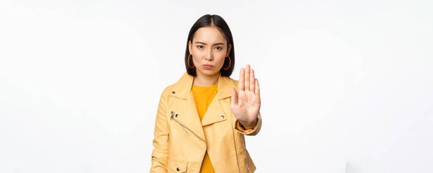 Ernstig en bezorgd aziatisch meisje uitbreiden palm hand, stoppen met gebaar, verbieden smth, verbieden, staande over witte studio achtergrond - Foto, afbeelding