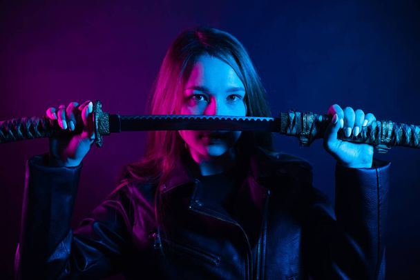 Cyber lány fekete bőrdzsekiben alkonyatkor egy katanát tart. Egy nő egy klubban színes rózsaszín-kék fénnyel egy ázsiai kardot tart. Harcos! - Fotó, kép