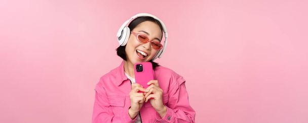 Tyylikäs aasialainen tyttö kuulokkeissa, musiikin kuunteleminen ja valokuvien ottaminen matkapuhelimella, älypuhelimella, vaaleanpunaisen taustan päällä - Valokuva, kuva