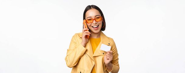 Tyylikäs aasialainen naismalli, puhuu älypuhelimella ja näyttää luottokortin, seisoo valkoisen taustan yläpuolella - Valokuva, kuva