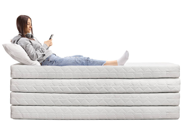 Feemale teenager on a pile of mattresses using a smartphone isolated on white background - Фото, изображение