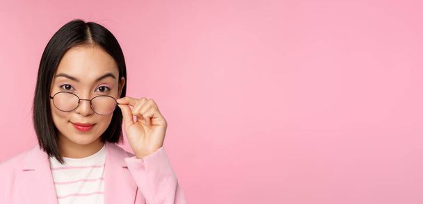 Portrait of asian businesswoman in glasses, looking intrigued at camera and smiling, professional saleswoman staring with interest, pink background - Fotoğraf, Görsel