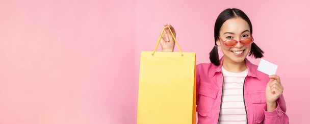幸せな若いアジアの女性は、クレジットカードやショッピングバッグを示しています,店舗販売のお知らせ,店でスミスを購入,ピンクを背景にポーズ - 写真・画像