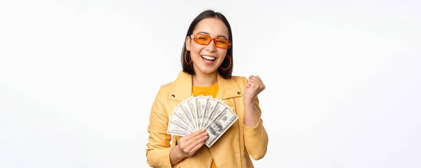 Стильна усміхнена азіатська дівчина тримає готівку, показуючи долари та святкуючи, стоїть на білому тлі
 - Фото, зображення