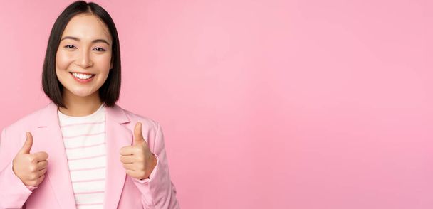 Επαγγελματική επιχειρηματίας, ασιατική εταιρική γυναίκα δείχνει αντίχειρες επάνω και χαμογελώντας, έπαινος και φιλοφρόνηση, στέκεται στο κοστούμι πάνω από ροζ φόντο - Φωτογραφία, εικόνα