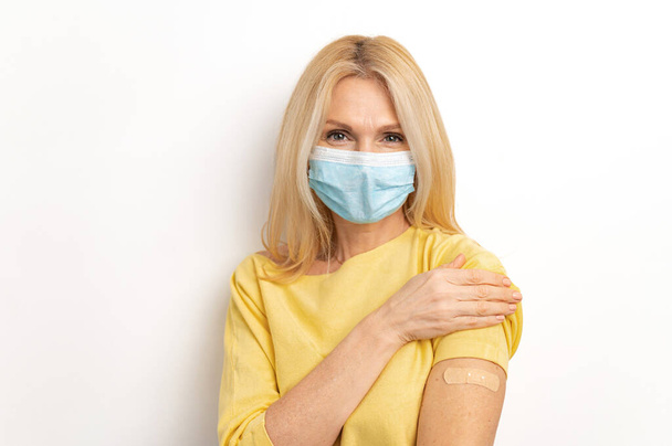 Portrait of caucasian senior woman wearing medical mask showing her arm with band aid after coronavirus Covid-19 vaccine - Zdjęcie, obraz