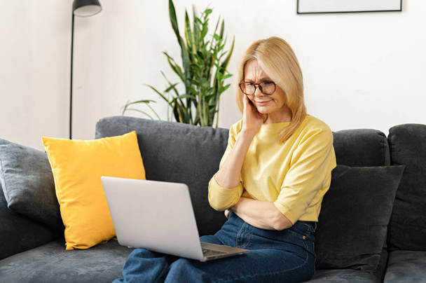 Brooding middle-aged woman using a laptop for remote work at home, studying online, a serious female mature teacher checking tasks - Foto, afbeelding