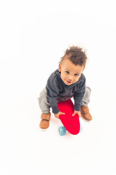Vertical studio shot of a cute innocent biracial baby boy looking at camera and holding red skateboard over white background. - Fotó, kép