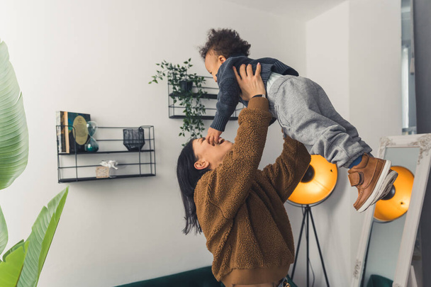 Young caucasian dark-haired mum lifting her biracial adorable son up in their modern flat. Relationship between parent and child. - Fotografie, Obrázek