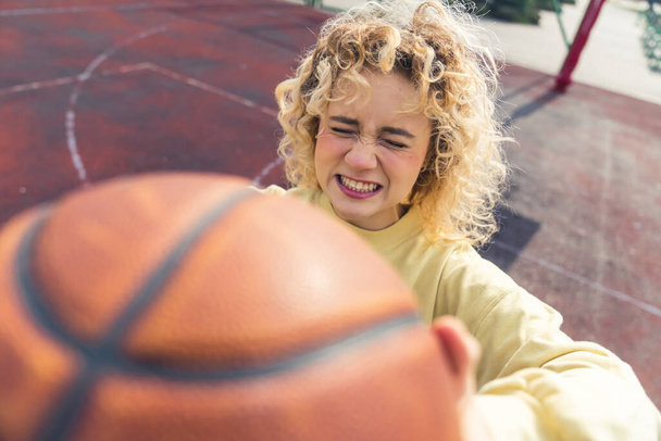 Młoda blond hipsterka pokazuje piłkę do koszykówki przed kamerą, robi głupią minę, stoi na boisku sportowym z bliska kopia przestrzeni  - Zdjęcie, obraz