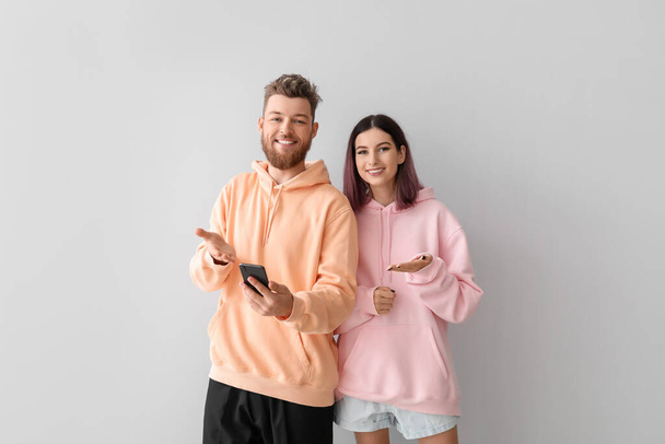 Cool young couple in hoodies and with phone on light background - Φωτογραφία, εικόνα