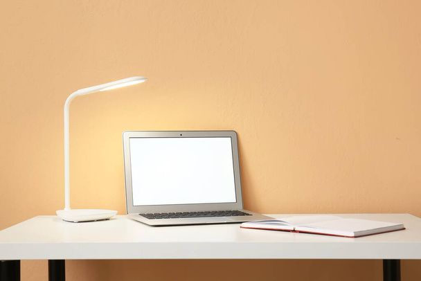 Lâmpada brilhante, laptop moderno e notebook aberto na mesa perto da parede de cor - Foto, Imagem
