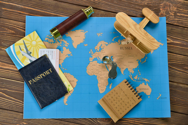 Passport with spyglass, airplane, calendar and world map on dark wooden background - Photo, Image