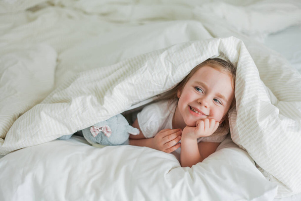 Pequeña linda chica rizada de ojos azules juega en la cama con su juguete de peluche. El niño está acostado en la cama debajo de las sábanas. Emociones. Sueño saludable. - Foto, Imagen