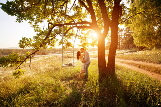 Jovem casal beijando sob grande árvore com balanço ao pôr do sol
 - Foto, Imagem