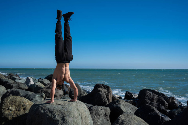 Shirtless man doing handstand on rocks by the sea.  - 写真・画像