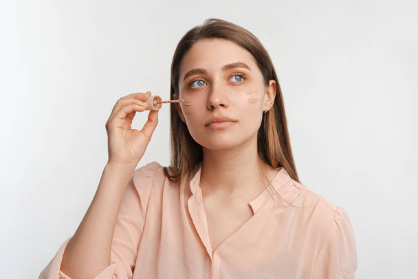 Young woman is applying cream foundation on her face while looking far away. - Photo, Image