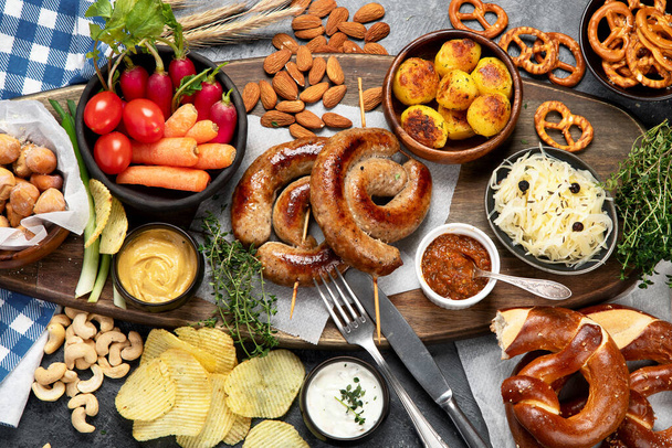 Beer and snack set. Oktoberfest food frame concept. Variety of  grilled sausages, fried potatoes, chips, vegetables, german pretzel pastry, almonds and sauces on dark wooden background. Top view. - Zdjęcie, obraz