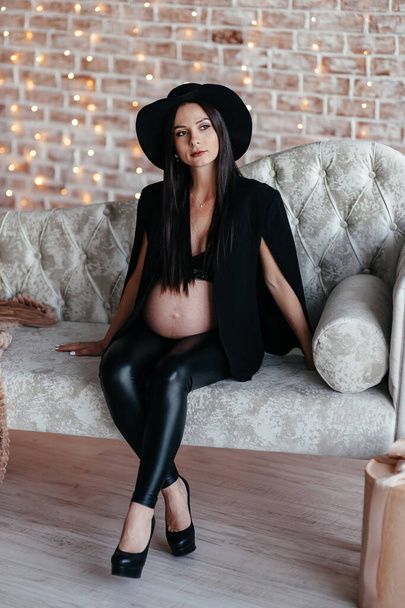 Young pregnant women ,long hair, wearing black dress and hat posing in studio.Grey background. - Фото, изображение