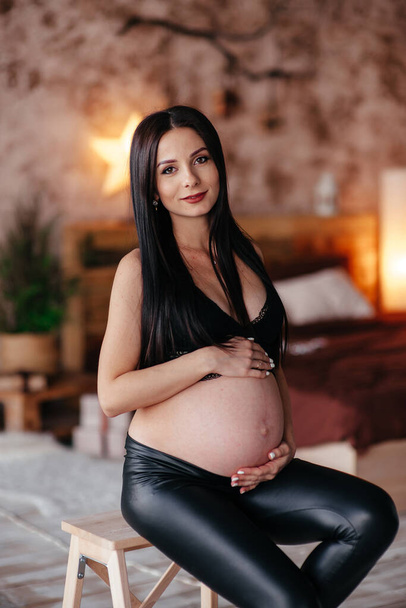 Fashionable pregnant mom, hugging belly. Stylish Motherhood concept. Happy young pregnant woman in stylish black bra and modern earring, holding belly bump and posing in light. fashion photo - Foto, afbeelding