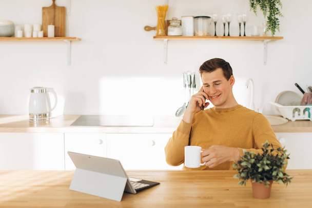 Красивый парень в желтой повседневной одежде сидит дома на кухне, пьет кофе и работает на планшете и телефоне - Фото, изображение