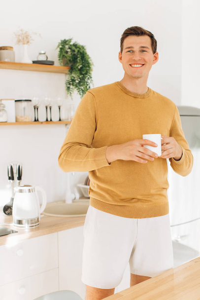 A handsome guy in yellow casual clothes is sitting at home in the kitchen drinking coffee - Foto, immagini