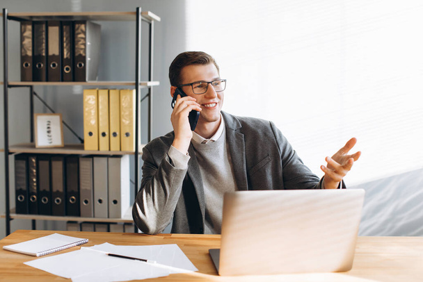 Современный человек в очках улыбается, офисный работник работает над ноутбуком и разговаривает по телефону - Фото, изображение