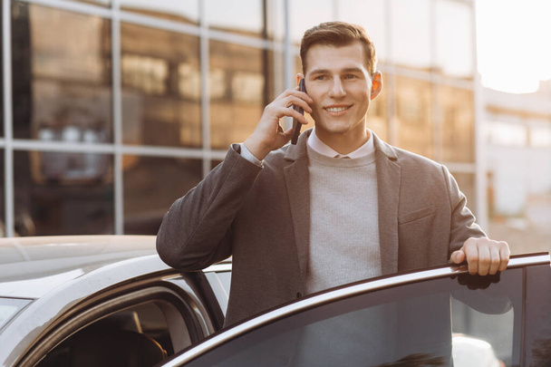 Όμορφος μοντέρνος νέος άντρας μιλάει τηλεφωνικά και μπαίνει στο αμάξι του ενώ στέκεται έξω. - Φωτογραφία, εικόνα