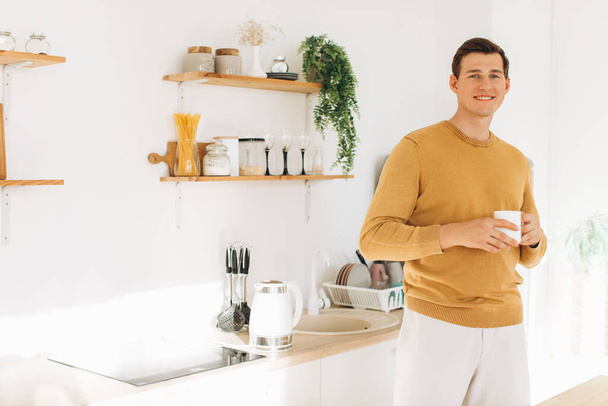 Ένας όμορφος άντρας με κίτρινα ρούχα κάθεται σπίτι στην κουζίνα πίνοντας καφέ. - Φωτογραφία, εικόνα