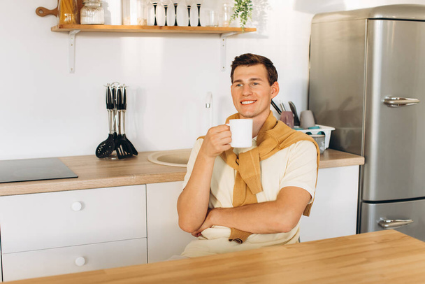 A handsome guy in yellow casual clothes is sitting at home in the kitchen drinking coffee - Valokuva, kuva