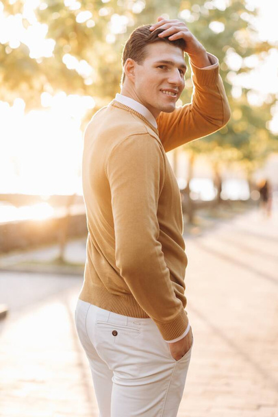 Modern handsome smiling man in yellow and white clothes posing in the park at sunset - Photo, image