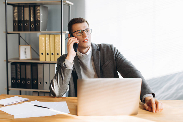 Homme moderne avec des lunettes souriant, employé de bureau travaillant sur ordinateur portable et ayant une conversation téléphonique - Photo, image