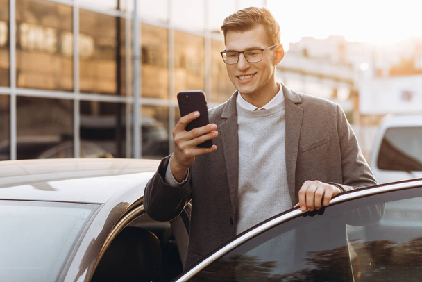 Schöner moderner junger Mann mit Brille, der telefoniert und im Freien in sein Auto steigt - Foto, Bild