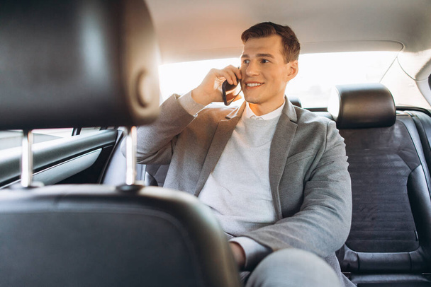 Jonge zakenman aan het praten op een mobiele telefoon terwijl hij op de achterbank van een auto zit. - Foto, afbeelding