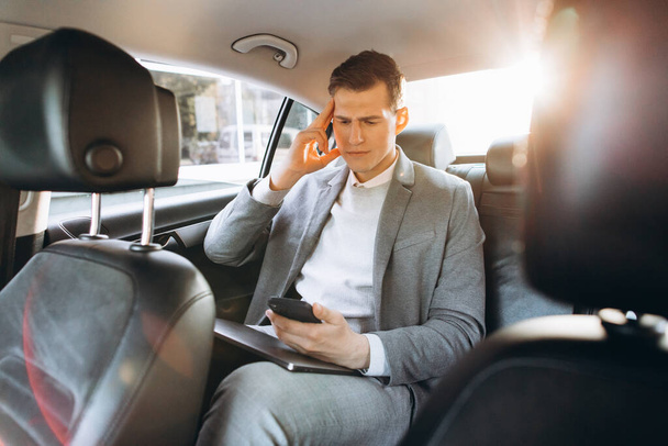 Mladý podnikatel unavený z práce drží ruku na hlavě, zatímco sedí na zadním sedadle auta. - Fotografie, Obrázek