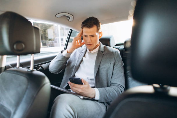 Junge Geschäftsmann müde von der Arbeit hält Hand an den Kopf, während er auf dem Rücksitz des Autos sitzt. - Foto, Bild