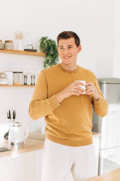 Un chico guapo con ropa casual amarilla está sentado en casa en la cocina bebiendo café. - Foto, imagen