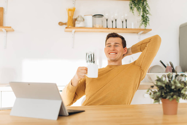 Knappe man in gele casual kleding zitten thuis in de keuken koffie drinken en werken op tablet en telefoon - Foto, afbeelding