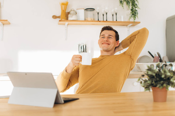 Handsome guy in yellow casual clothes sitting at home in the kitchen drinking coffee and working on tablet and phone - Фото, зображення